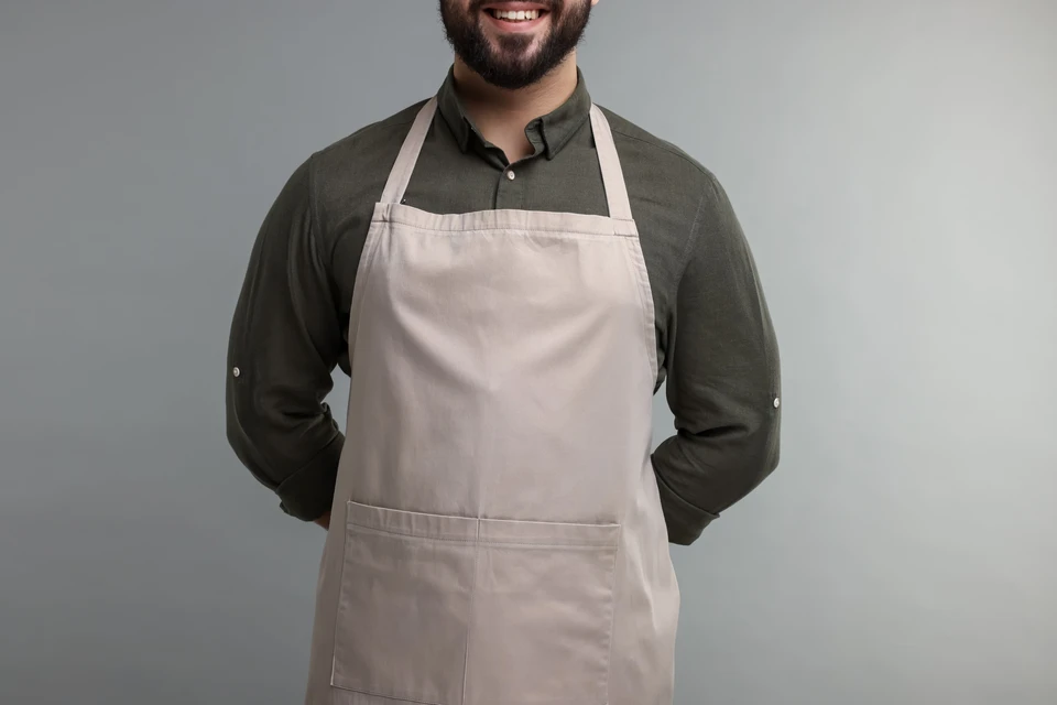 Homme en tenue professionnelle dans un environnement neutre, portant un tablier de cuisine beige clair par-dessus une chemise olive à manches longues