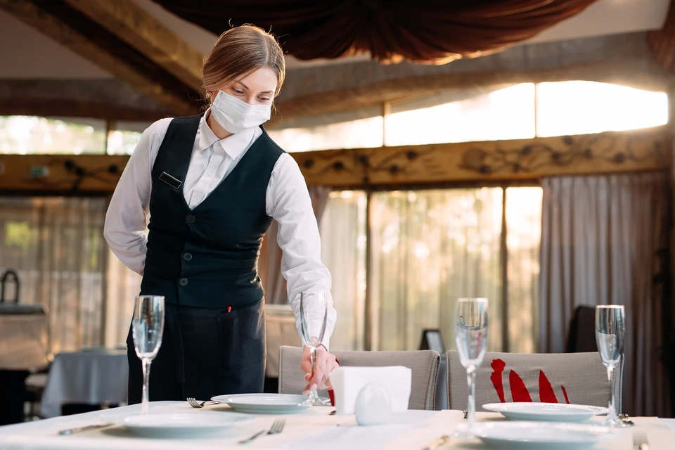 Serveuse en uniforme professionnel (gilet noir, chemise blanche) portant un masque sanitaire dresse une table de restaurant