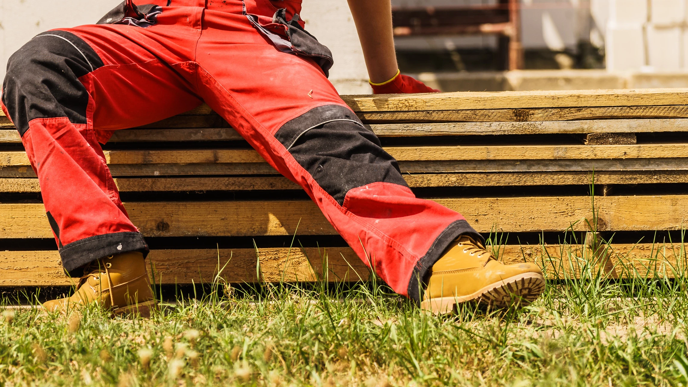 Ouvrier en combinaison rouge et noire assis sur palette en bois, portant chaussures de sécurité beiges